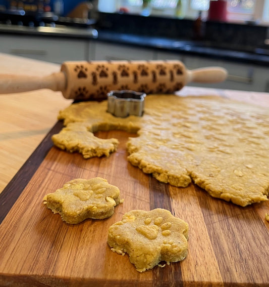 A wooden rolling pin with paw prints design, a metal cookie cutter, and rolled out dough on a wooden cutting board shaped into paw print and bone forms for dog treats.